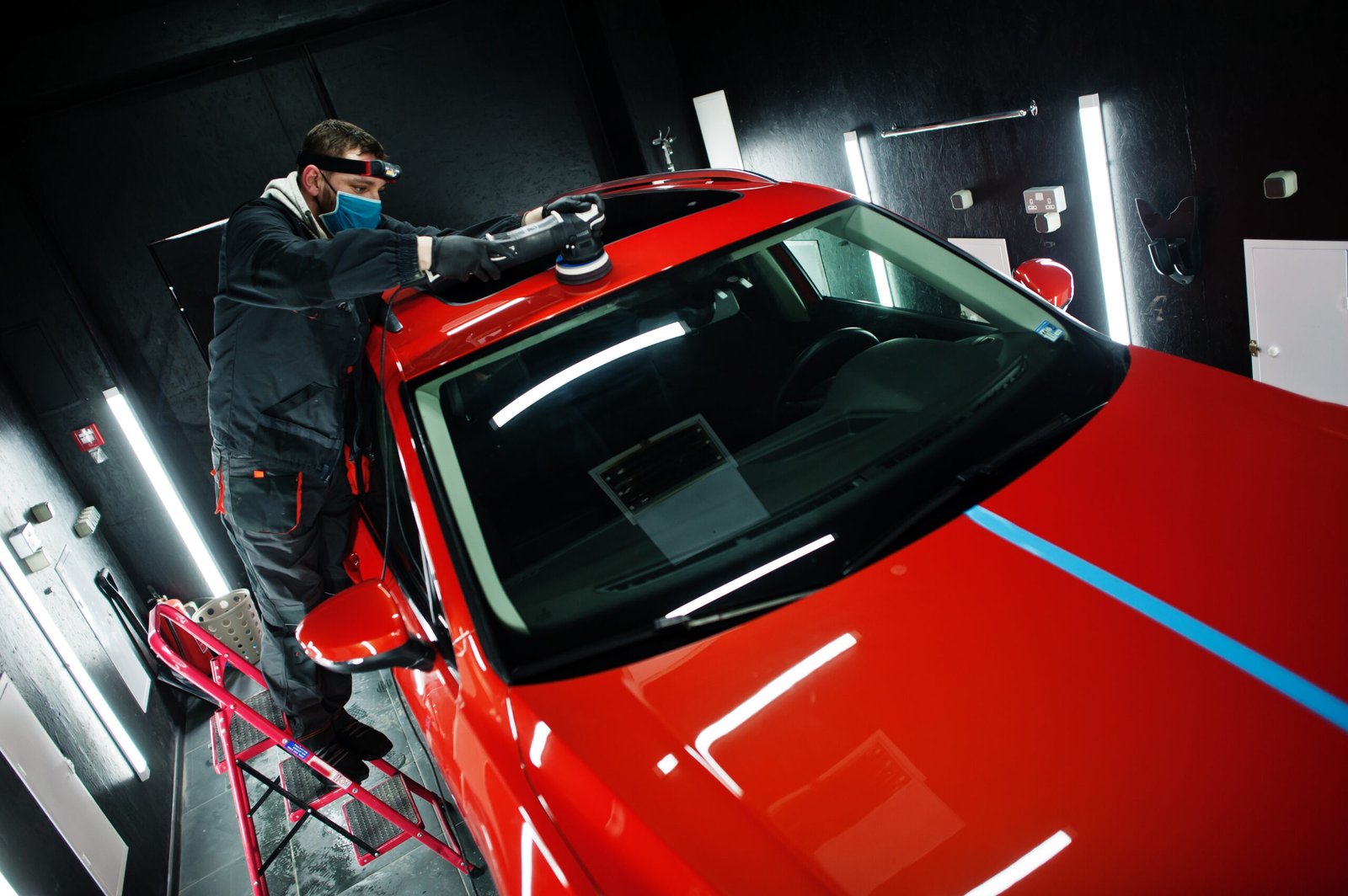 Car detailing concept. Man in face mask with orbital polisher in repair shop polishing roof of orange suv car.